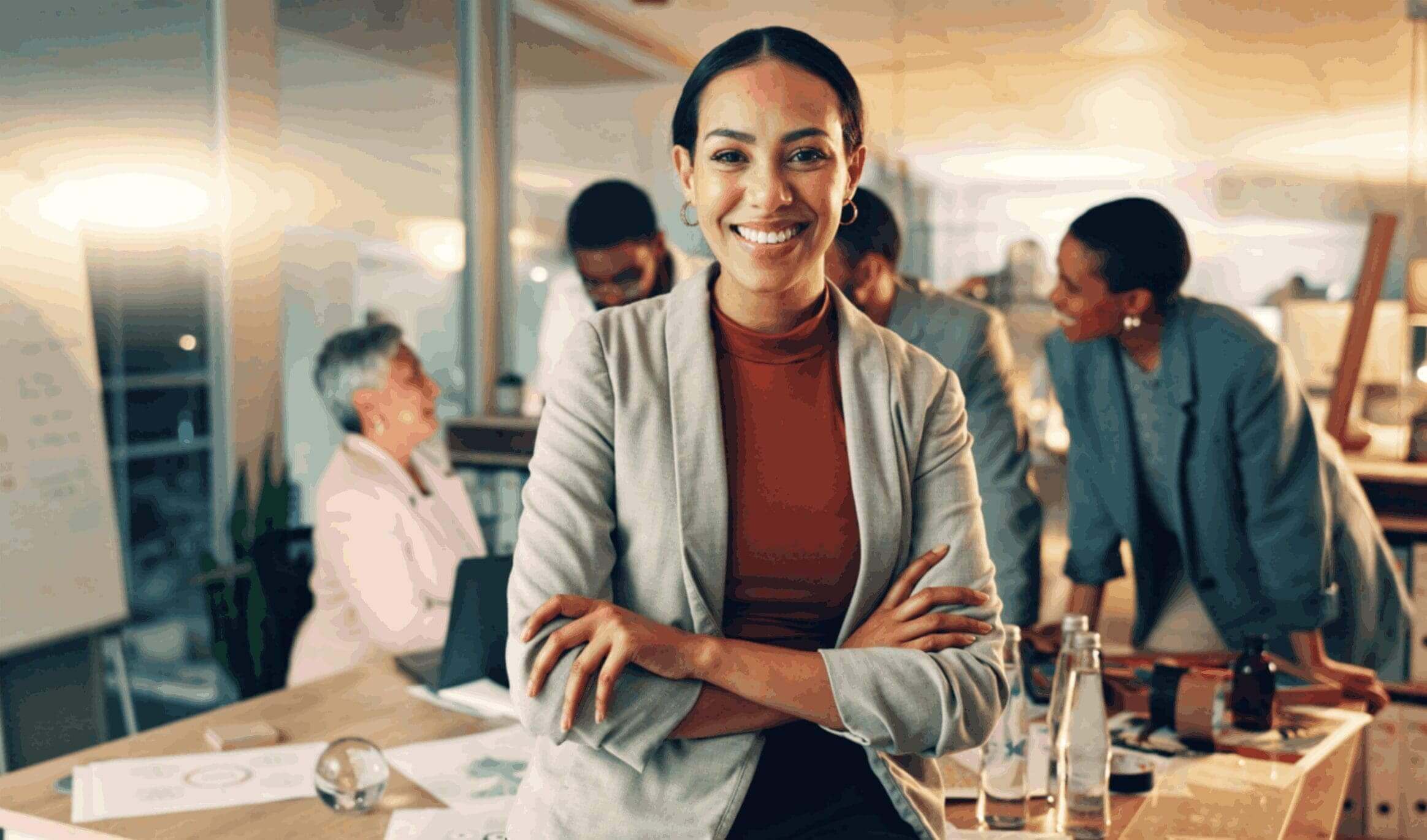 Confident woman smiling with arms crossed in a busy office.